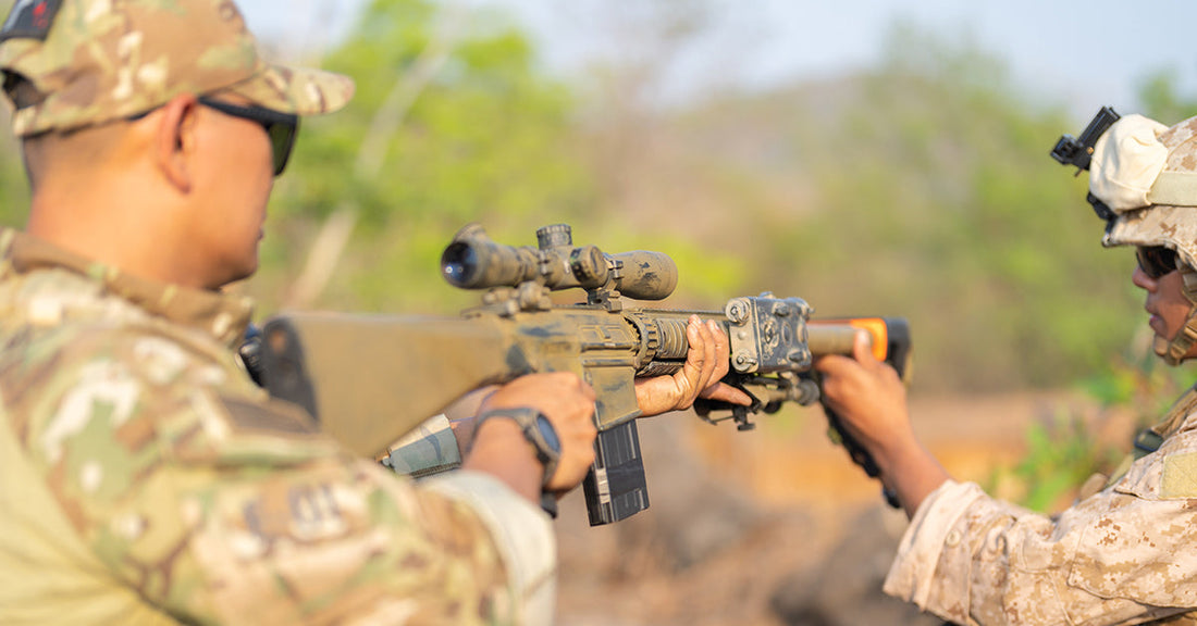 Two soldiers in camouflage uniforms participate in a weapon-handling drill outdoors, one holding a rifle steady while the other assists with positioning.
