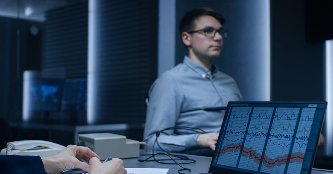 Examiner monitoring a laptop displaying polygraph charts while a man with sensors on his chest and arm sits in the background.