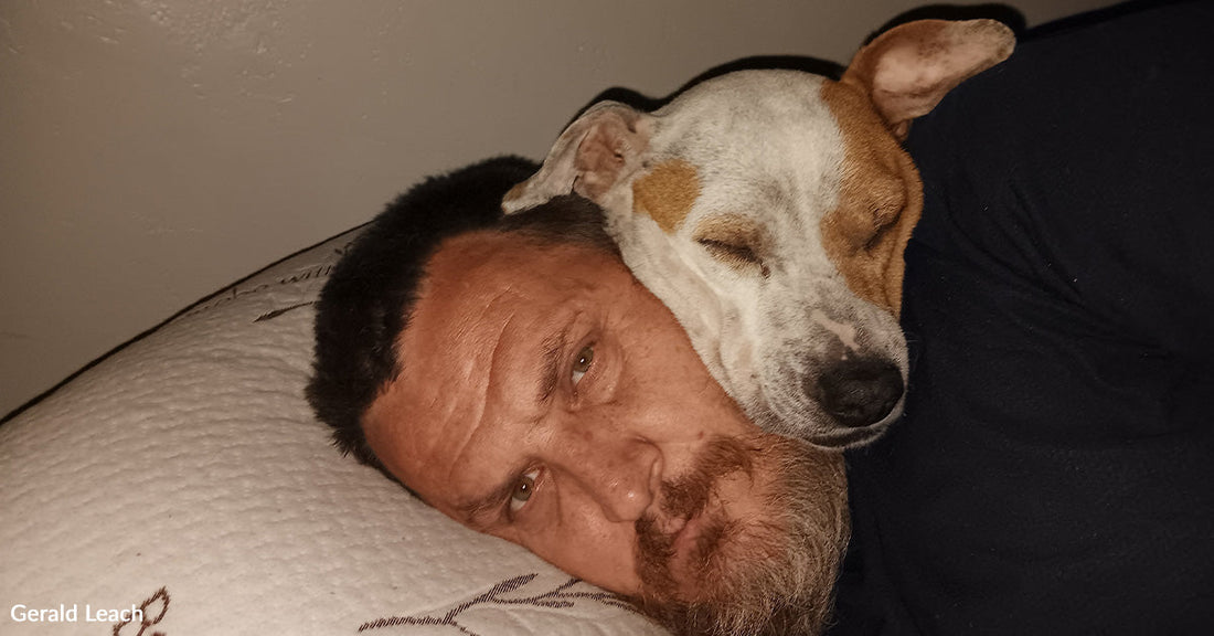 A man lies on a pillow while a small white-and-brown dog sleeps with its head resting on his face, both appearing calm and relaxed.