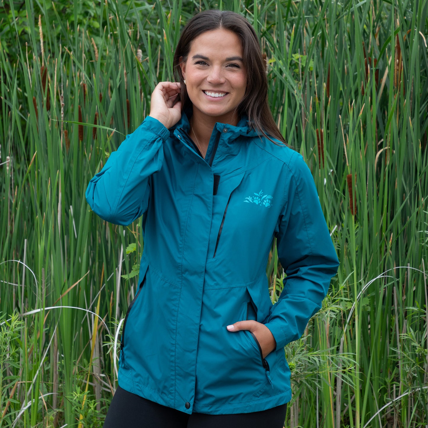Woman wearing a teal rain jacket standing in tall green grass.