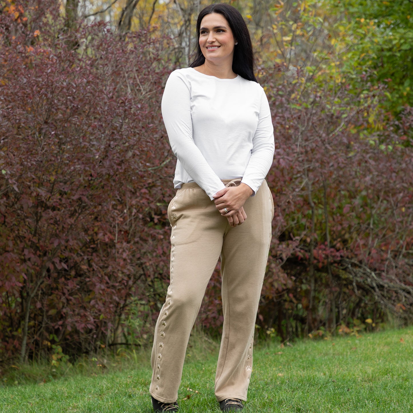 Woman wearing a white long-sleeve top and beige pants standing in a grassy area with trees in the background.