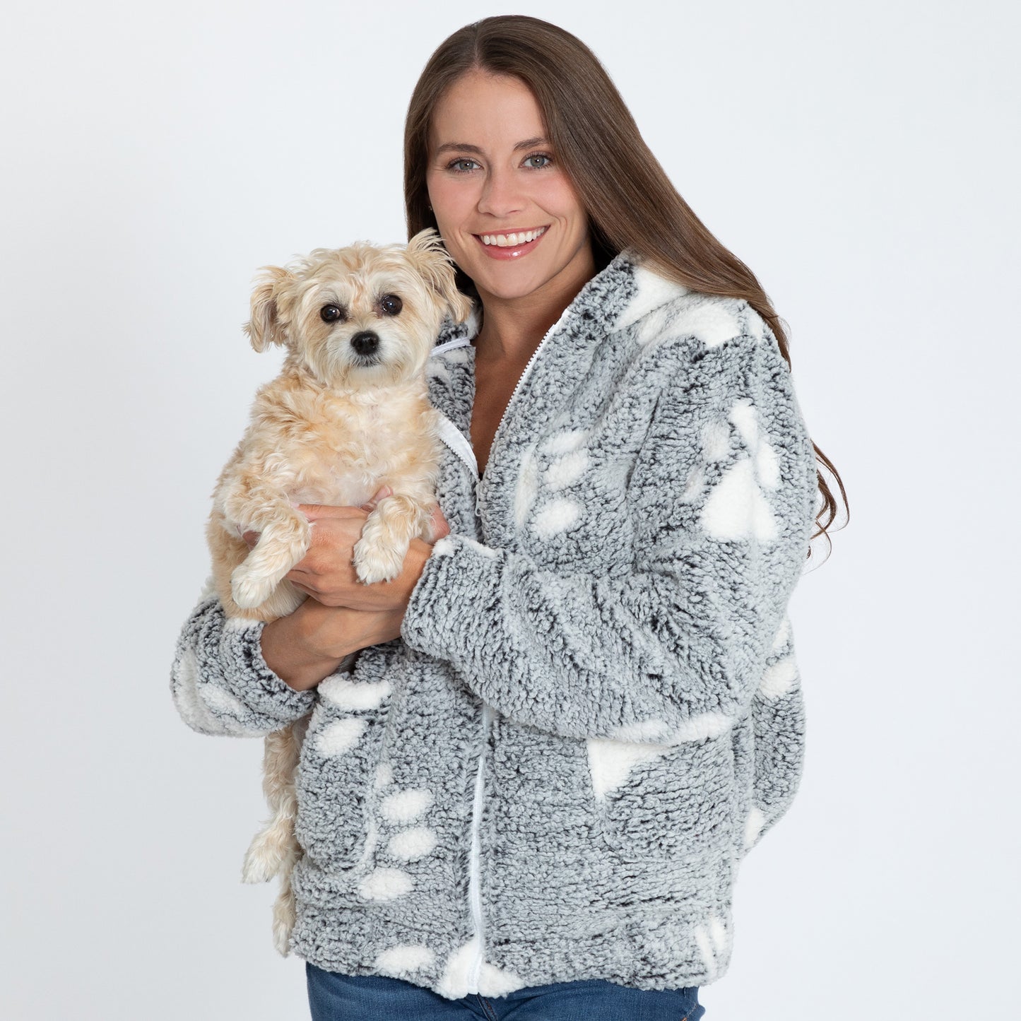 Woman holding a small dog, wearing a gray sherpa fleece jacket with white paw prints, with two front pockets, a hood, and a full zip front with a white background.