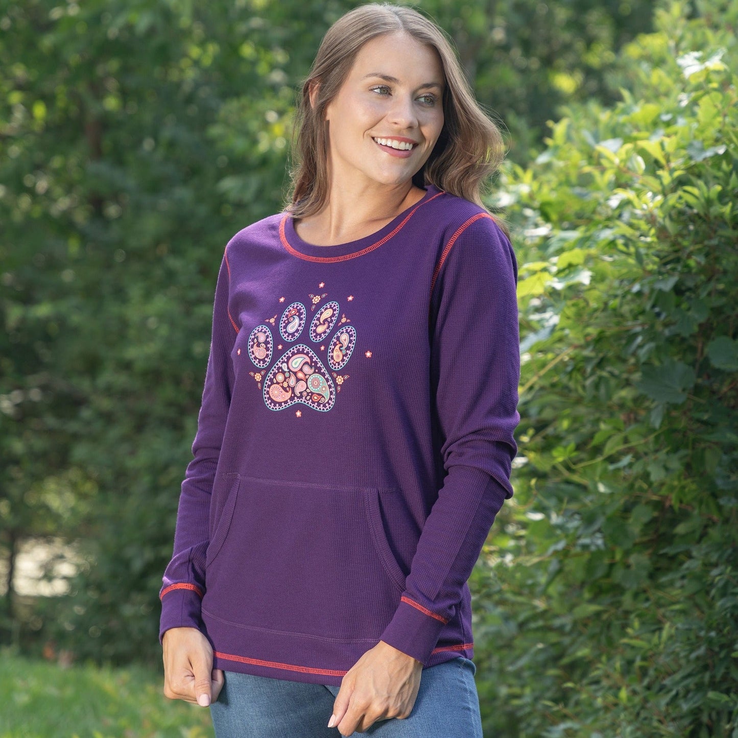 Woman wearing a purple thermal long sleeve top with a kangaroo pocket, and a paisley paw print design on the front, in a tree filled outdoor setting.