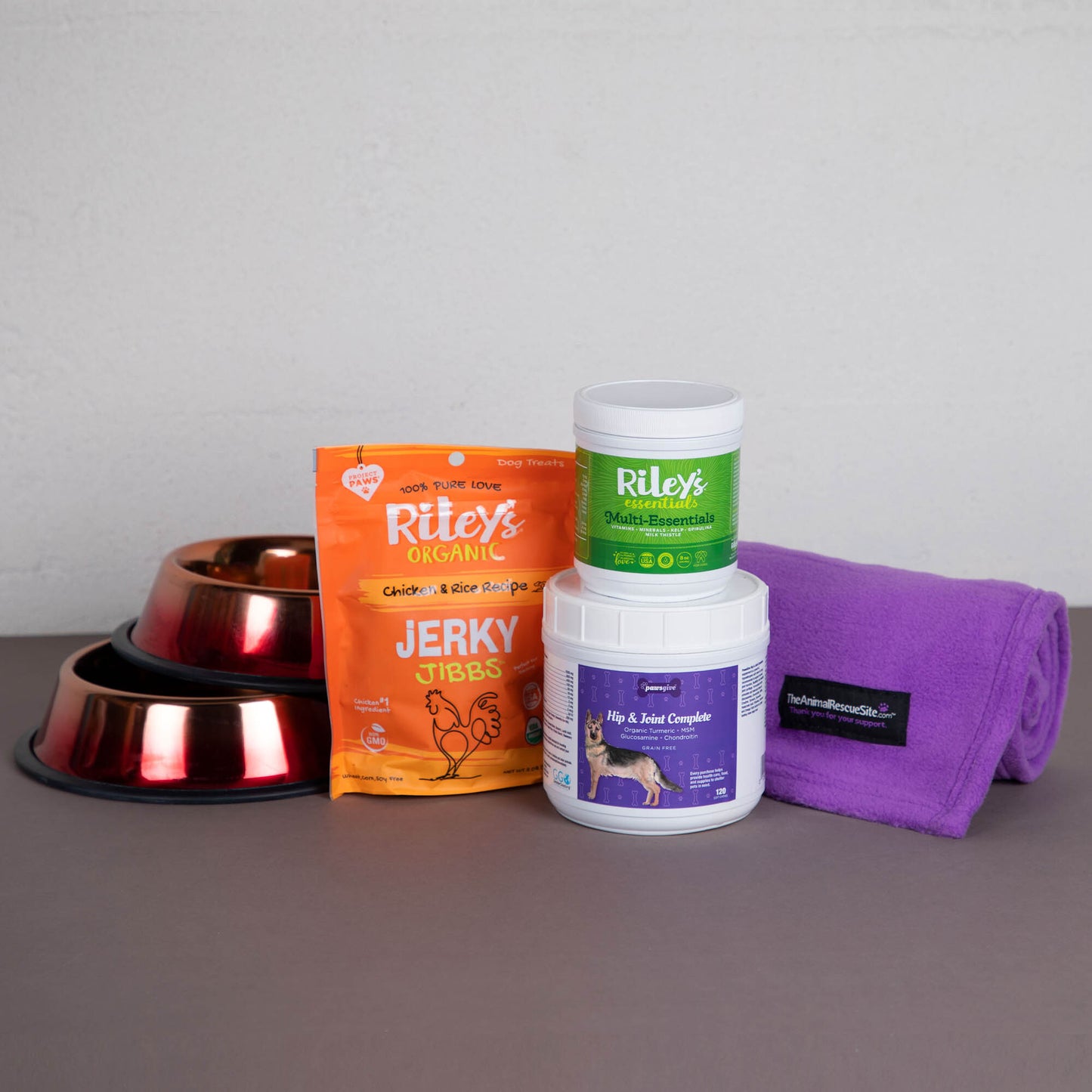 Two bowls, bag of dog treats, joint supplements, essential vitamin powder and a purple blanket.