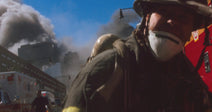 An FDNY firefighter wearing a dust mask and helmet pulls a hose beside a fire truck amid billowing smoke in a city street.