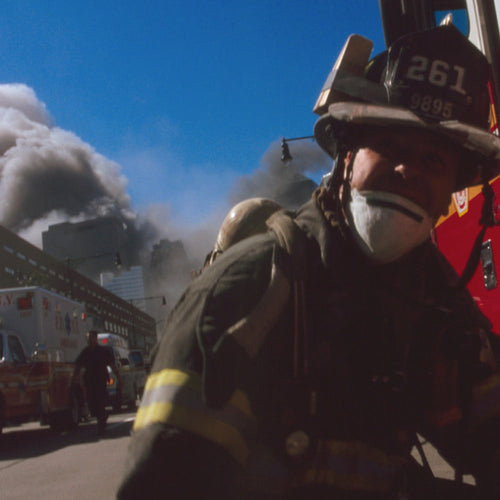 An FDNY firefighter wearing a dust mask and helmet pulls a hose beside a fire truck amid billowing smoke in a city street.