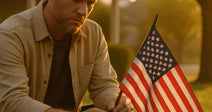 Man kneels on a front lawn at sunset, holding a small American flag outside a suburban home.