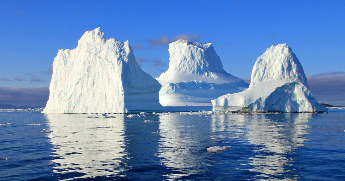 Towering Antarctic icebergs reflect off still ocean water under a clear blue sky.