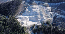 An aerial view of a large open-pit quarry carved into a forested mountainside, with snow-capped peaks visible in the distance.