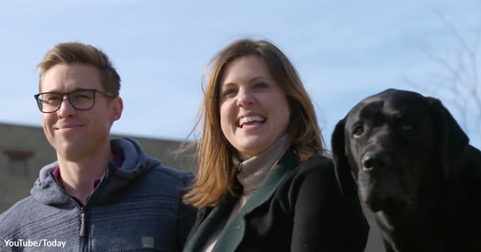 Two adults stand outdoors beside a black Labrador, all facing forward under a clear blue sky.