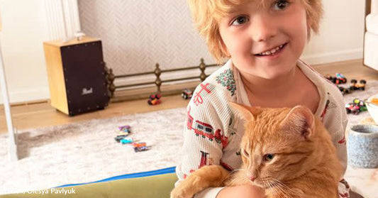 Blond child indoors smiles while holding an orange tabby cat in their arms.