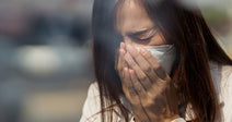 Woman wearing a face mask outdoors, covering her nose and mouth with both hands as if reacting to poor air quality.