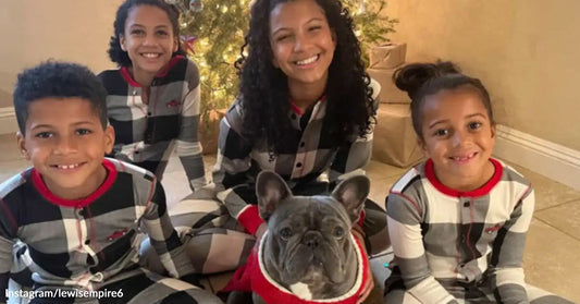 Four children wearing matching pajamas sit smiling beside a gray French bulldog dressed in a red sweater in front of a Christmas tree.