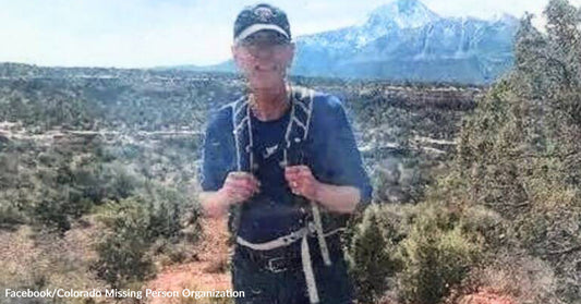 Man wearing a cap and backpack standing on a hiking trail with scrubland and snow-capped mountains in the background.
