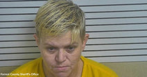 Mugshot of a woman with short blond hair wearing a yellow shirt in front of booking height lines.