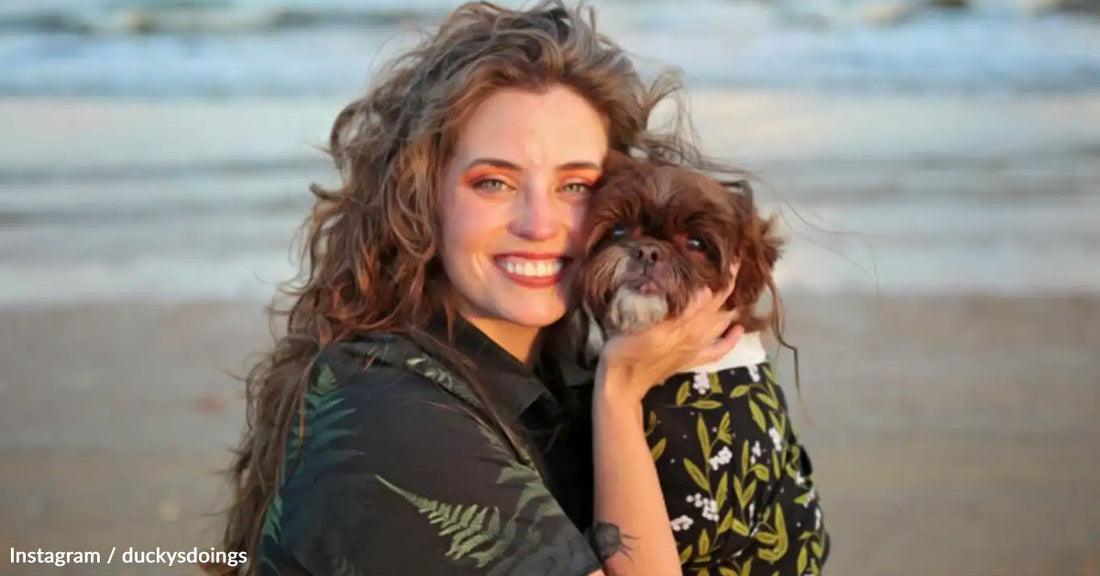 Smiling woman holds a small dog by the beach at sunset.