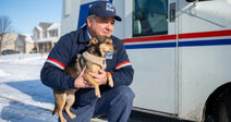 A postal worker kneels in the snow, holding a small dog outside a delivery truck.