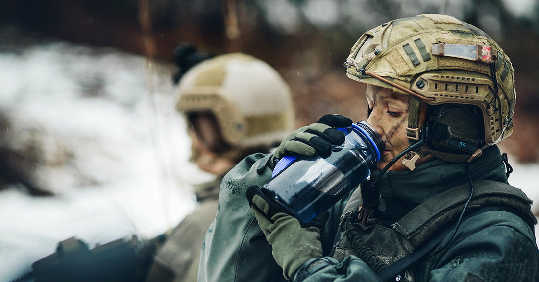 Soldier in combat gear and helmet drinks from a blue water bottle while resting in a cold outdoor environment.