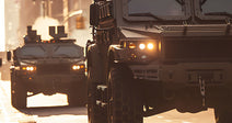 Armored military vehicles drive down a city street in formation, their headlights on as smoke rises in the background.