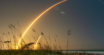 Long-exposure image of a rocket launching near a beach at dusk, with bright curved light trails arcing across the sky above sea grass and shoreline.