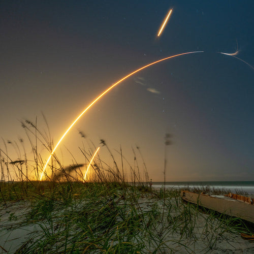 Long-exposure image of a rocket launching near a beach at dusk, with bright curved light trails arcing across the sky above sea grass and shoreline.
