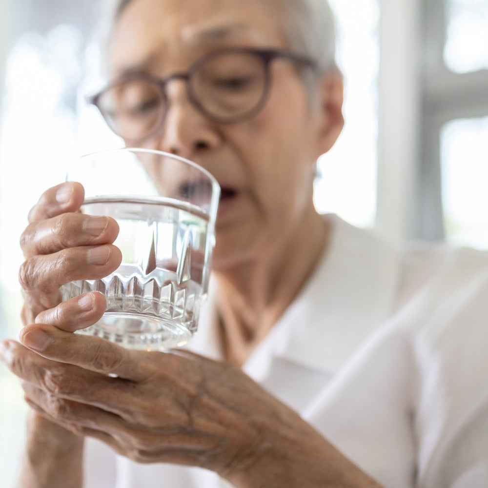 An elderly woman with white hair and glasses holds a glass of water up to her lips near a bright window.