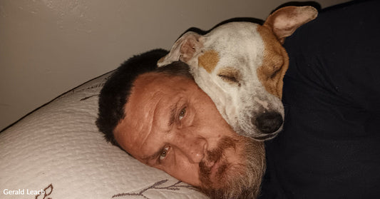 A man lies on a pillow while a small white-and-brown dog sleeps with its head resting on his face, both appearing calm and relaxed.