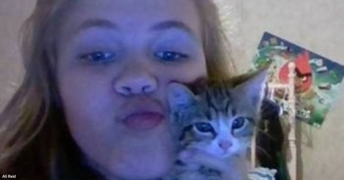A young woman with blonde hair holds a small tabby kitten close to her cheek while making a kissing face indoors.
