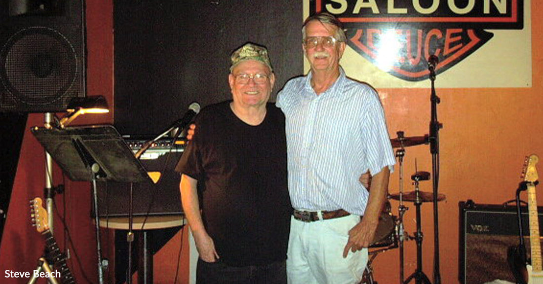 Two older men standing arm in arm on a small stage with guitars and drums behind them, beneath a “Double Saloon” sign.