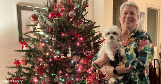 A woman stands beside a decorated Christmas tree holding a small white dog, both facing the camera in a warmly lit living room.