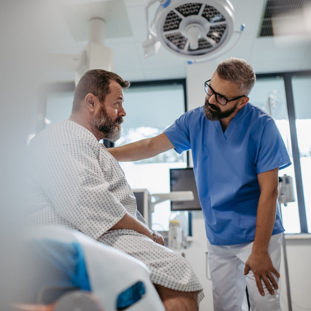 A healthcare worker in blue scrubs places a reassuring hand on a seated patient's shoulder in a modern medical examination room.