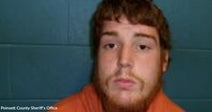 Mugshot of a young man with short reddish hair and beard wearing an orange jail shirt.