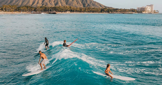 Surfer lying flat on a board, paddling toward the horizon as the sun sets over glowing golden water.