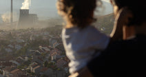 A woman holding a small child looks out over a town blanketed in haze, with a smoking industrial plant in the distance against mountain slopes.