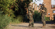 Two soldiers in camouflage uniforms and tactical gear stand outdoors, one holding a rifle while the other drinks from a water bottle near an abandoned building.