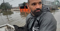 Man in a rain jacket rides through deep floodwater in a small boat while a black dog sits in front of him, with flooded streets and buildings in the background.