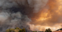 Massive dark smoke clouds from a wildfire billow above a suburban neighborhood of tiled-roof homes and green trees.