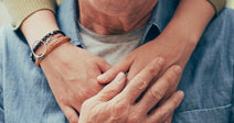 An elderly man sitting indoors is warmly embraced from behind by a younger person resting their arms across his chest.