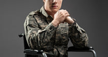 A young man in military camouflage uniform sits in a wheelchair against a gray studio background, hands clasped together.