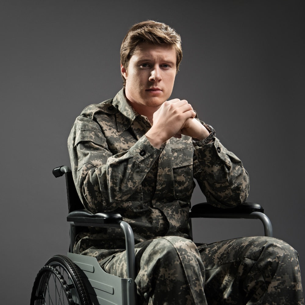 A young man in military camouflage uniform sits in a wheelchair against a gray studio background, hands clasped together.