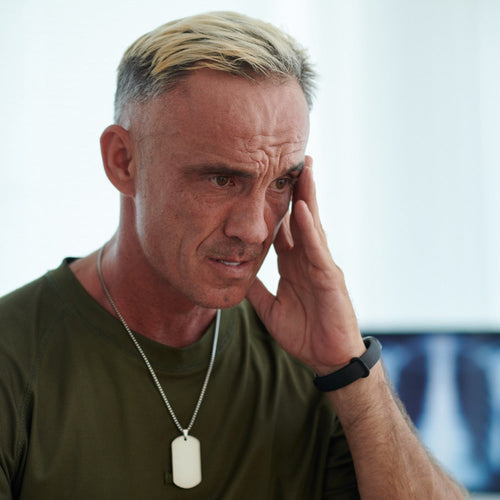 A middle-aged man wearing a military dog tag and olive green shirt holds his hand to his temple with a distressed expression, seated in a bright medical office.