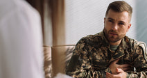 Man in military camouflage uniform sitting in a counseling session, holding his chest and speaking seriously while a clinician takes notes nearby.