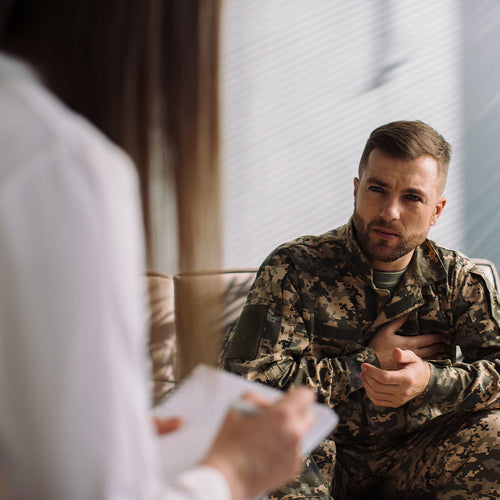 Man in military camouflage uniform sitting in a counseling session, holding his chest and speaking seriously while a clinician takes notes nearby.