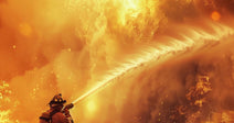 Firefighter in protective gear sprays a powerful stream of water toward a towering wall of flames and smoke.
