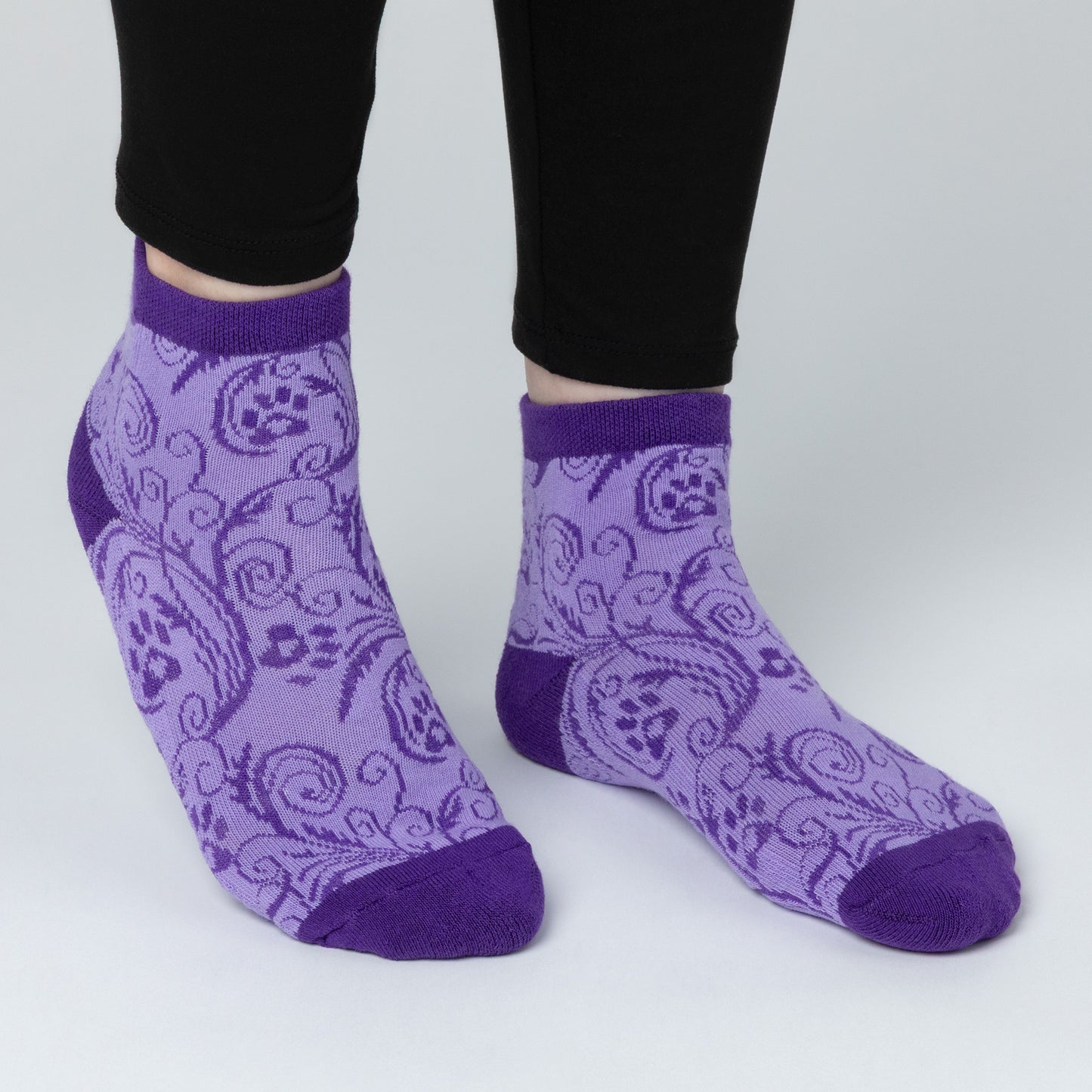 A person wearing ankle length comfortable cotton-blend socks that are lavender color contrasted with a dark purple paw print and swirl design. The cuff, heels and toes of the sock are a plain dark purple. 