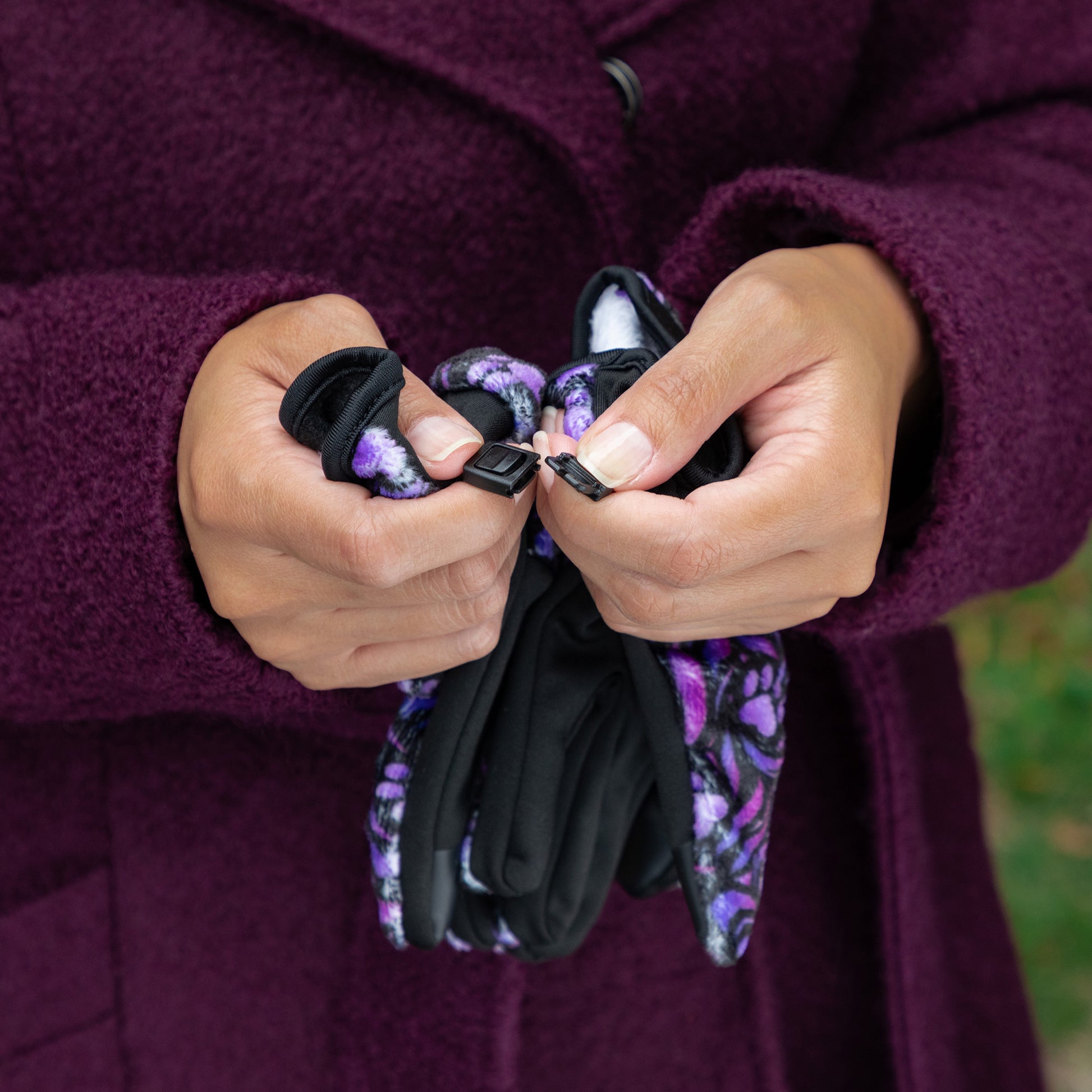 A woman wearing a coat unclasps a pair of touchscreen technology fingertips warm black fleece gloves adorned with purple paw prints and swirls. 