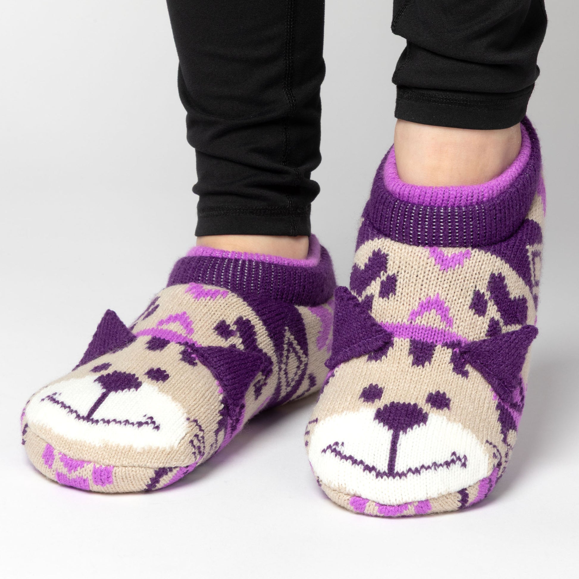 Person wearing purple and beige patterned knitted slipper socks with a smiling cat face on top, on a white background beige cat