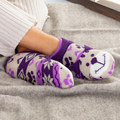 Person wearing purple and beige patterned knitted slipper socks with a smiling cat face on top, on a gray blanket beige cat