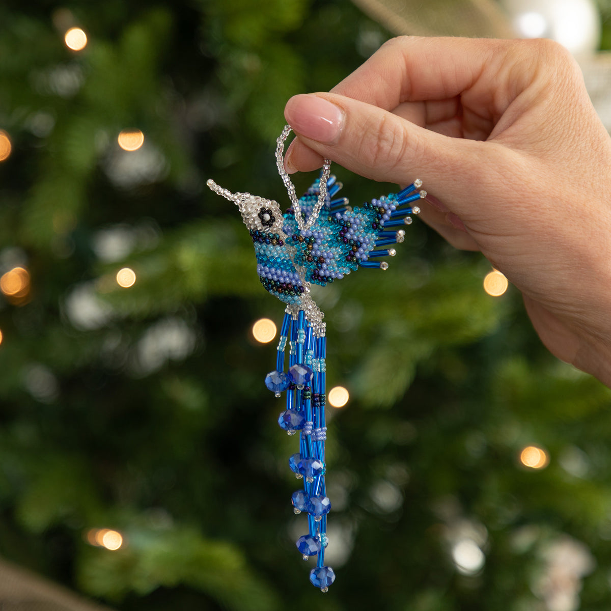 A hand holds a beaded bird Christmas ornament with a long blue tail, set against a blurred green Christmas tree with warm lights.