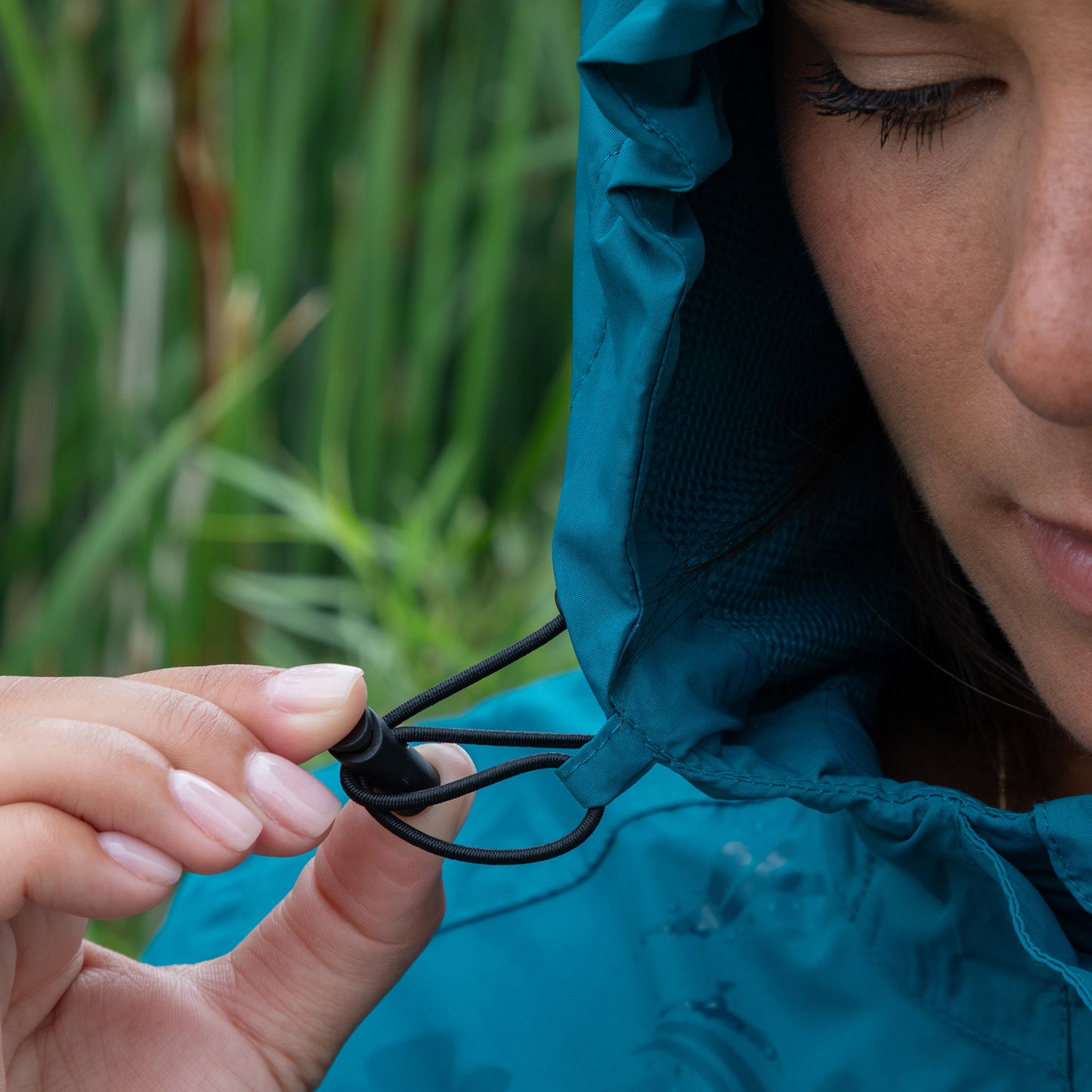 Person adjusting the hood of a blue raincoat with a blurred green background.